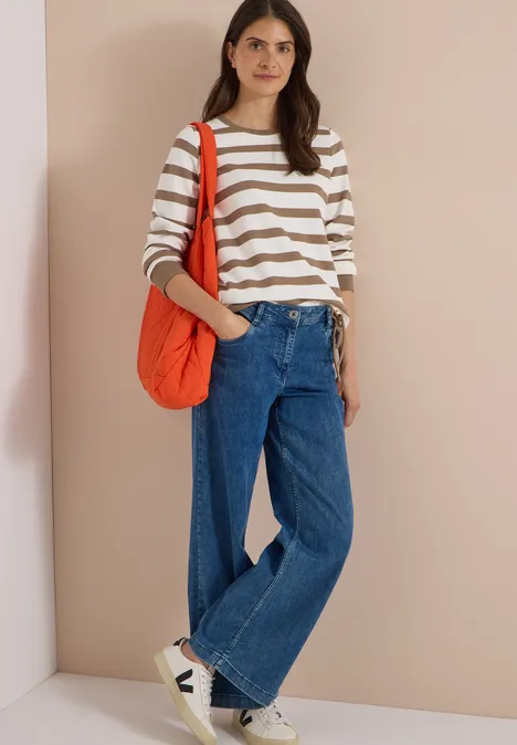 Eine Frau in weiß-beige gestreiftem Pullover, weitem Jeans, weißen Sneakern und orangener Tasche steht vor einer Wand.