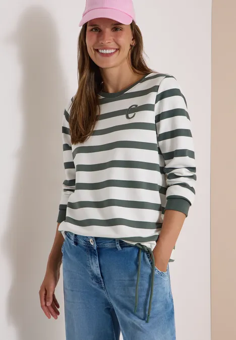 Frau lächelnd in rosa Cap, weiß-grün gestreiftem Shirt und lockeren Jeans.