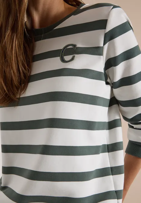 Eine Person trägt ein weiß-grün gestreiftes 3/4-Arm-Shirt mit rundem Ausschnitt und dezentem Logo.