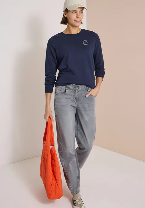 Frau in dunkelblauem Sweatshirt, grauen Jeans und Cap, hält eine orange Stepp-Tasche und trägt Sneakers.