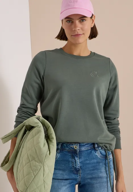 Junge Frau mit rosa Cap trägt grünes Sweatshirt, Jeans und hält eine hellgrüne Steppjacke vor neutralem Hintergrund.