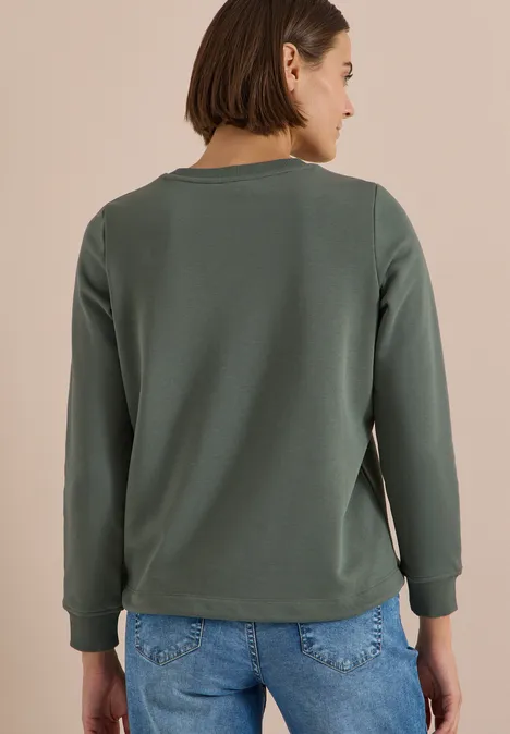 Frau steht Rücken zur Kamera, trägt grünes Sweatshirt und blaue Jeans.