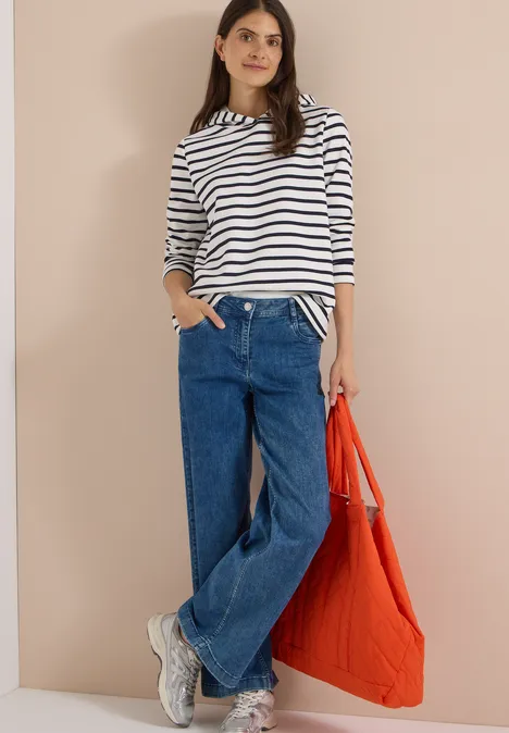 Sweatshirt mit Kapuze und Streifen vanilla white Frau in gestreiftem Langarmshirt, weitem Jeans, weißen Sneakers und orangefarbener Tasche vor neutralem Hintergrund.