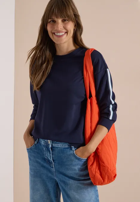 Sweatshirt mit Rundhals und Tapedetails urban dark blue Frau in Jeans, dunkelblauem Sweatshirt mit weißen Streifen und oranger Tasche lächelt.
