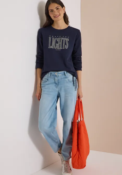 Sweatshirt mit Wording und Tunnelzug urban dark blue Eine Frau lehnt an einer Wand, trägt ein dunkelblaues Shirt mit Schriftzug, helle Jeans und hält eine orange Tasche.