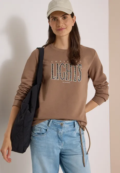 Sweatshirt mit Wording und Tunnelzug creamy mocha Junge Frau mit beigefarbener Cap, braunem Sweatshirt mit Schriftzug LIGHTS, Jeans und schwarzer Tasche.