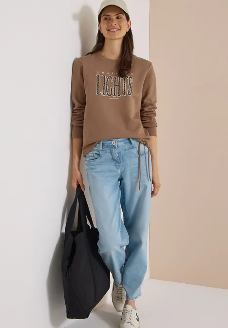 Sweatshirt mit Wording und Tunnelzug creamy mocha Frau in Jeans, braunem Pullover mit Aufdruck 'LIGHTS', Cap und schwarzer Tasche lehnt an weißer Wand.