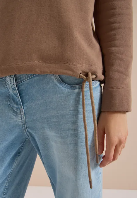 Sweatshirt mit Wording und Tunnelzug creamy mocha Eine Person trägt eine braune Pulloverjacke mit Tunnelzug und hellblaue Jeans; die linke Hand ruht locker an der Seite.