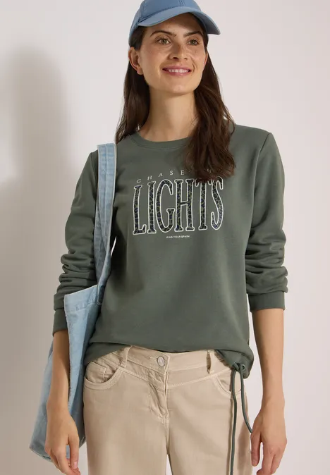 Sweatshirt mit Wording und Tunnelzug dried olive green Frau lächelt, trägt ein olivgrünes Sweatshirt mit Aufschrift 'LIGHTS', helle Hose, blaue Cap und Tasche.