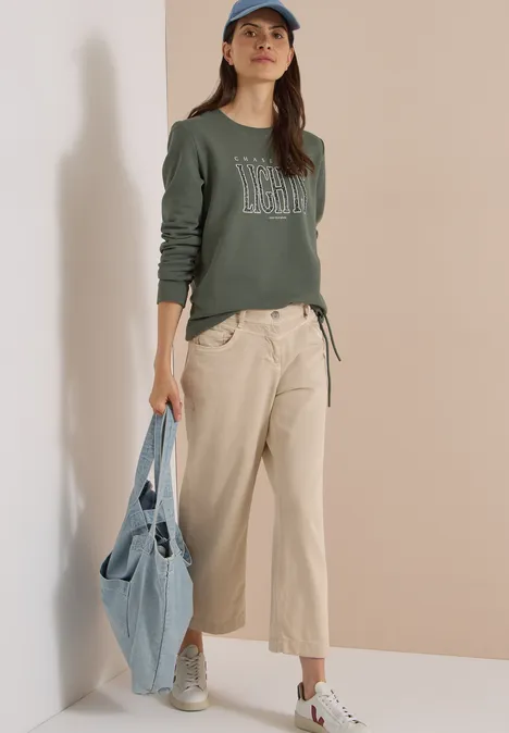 Sweatshirt mit Wording und Tunnelzug dried olive green Frau in grünem Sweatshirt, beigen Hosen, weißen Sneakern und Baseballcap, hält eine Jeans-Tasche.