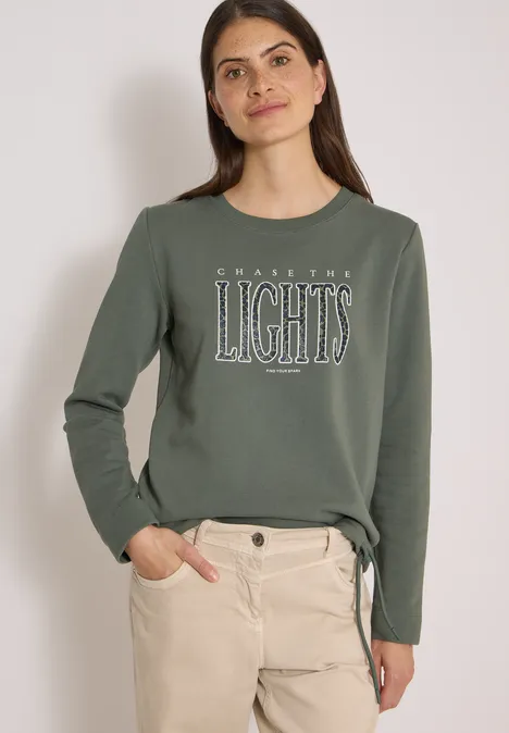 Sweatshirt mit Wording und Tunnelzug dried olive green Frau in olivgrünem Sweatshirt mit Aufdruck 'LIGHTS' und hellen, hochgekrempelten Hosen, lässig stehend.