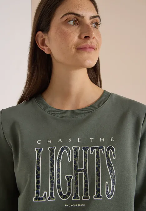 Sweatshirt mit Wording und Tunnelzug dried olive green Frau mit langen dunklen Haaren trägt grünes Shirt mit Aufschrift 'CHASE THE LIGHTS'. Sie blickt seitlich.