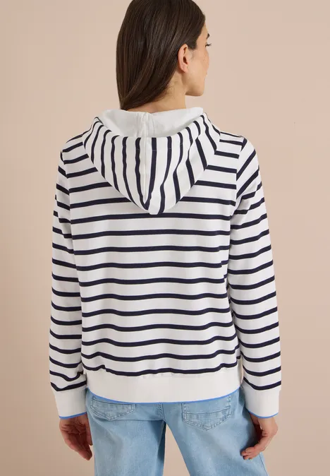 Frau von hinten in weißer Kapuzenjacke mit dunkelblauen Streifen und blauen Jeans.