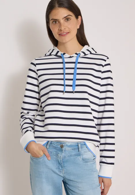 Junge Frau trägt einen weiß-blau gestreiften Kapuzenpullover und Jeans vor hellem Hintergrund.