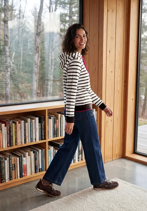 Sweat à capuche rayé truffle brown Une femme en pull rayé marche dans une pièce lumineuse avec une bibliothèque et de grandes fenêtres.