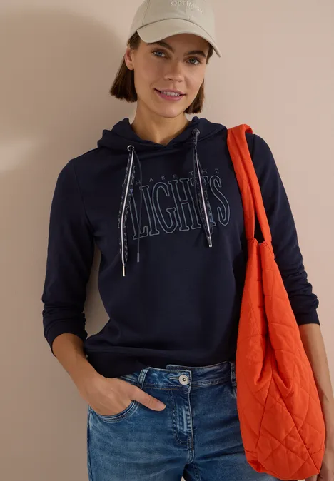 Sweatshirt mit Kapuze und Wording urban dark blue Frau mit Baseballcap trägt dunkelblauen Hoodie mit Schriftzug, Jeans und orangefarbene Schultertasche.
