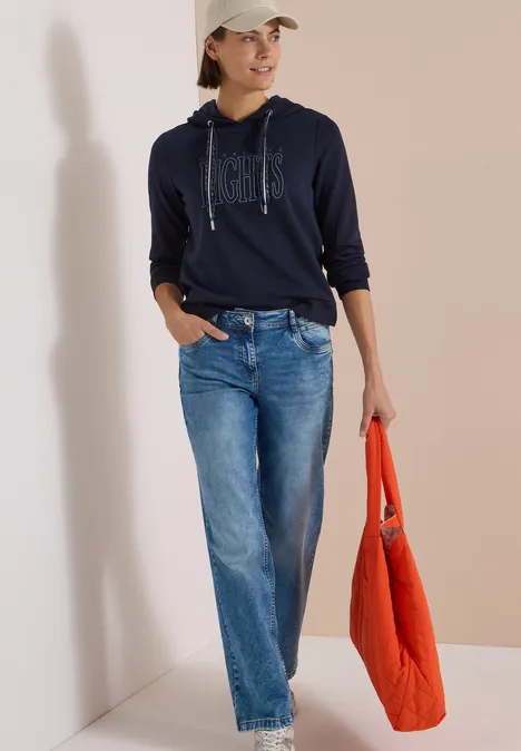 Sweatshirt mit Kapuze und Wording urban dark blue Frau in Navy-Hoodie, weiten Jeans und Basecap, trägt eine orangefarbene Tasche.