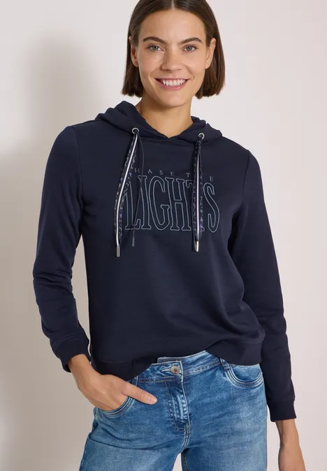 Sweatshirt mit Kapuze und Wording urban dark blue Frau in dunkelblauem Hoodie mit 'NIGHTS'-Aufdruck und Jeans lächelt entspannt in die Kamera.