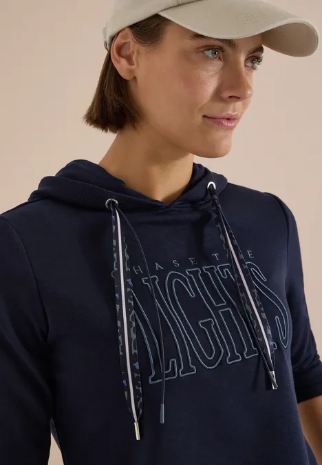 Sweatshirt mit Kapuze und Wording urban dark blue Frau in dunkelblauem Hoodie mit modischem Schriftzug und heller Cap, Nahaufnahme.