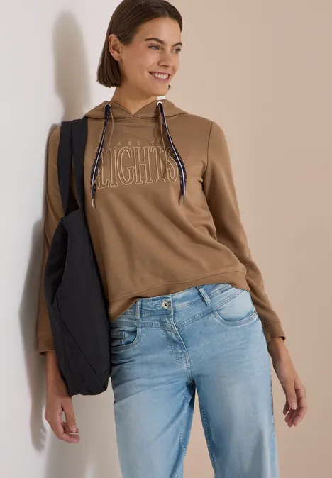 Sweatshirt mit Kapuze und Wording creamy mocha Junge Frau lehnt lächelnd an Wand, trägt braunen Hoodie, hellblaue Jeans und schwarzen Beutel.