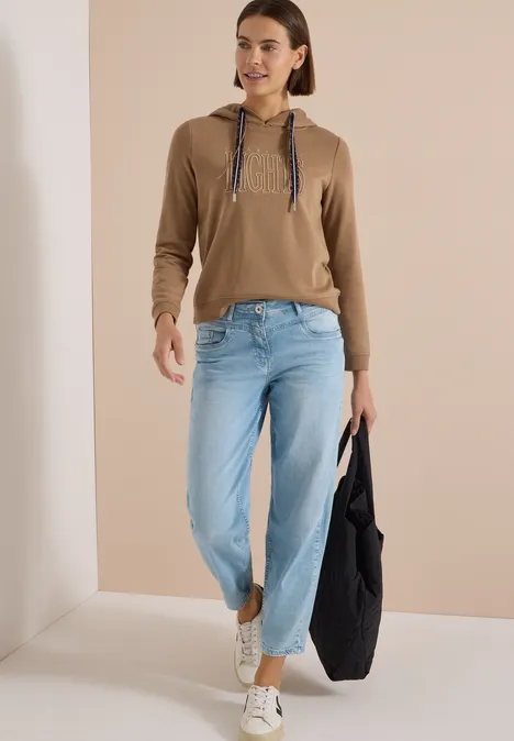 Sweatshirt mit Kapuze und Wording creamy mocha Frau in brauner Kapuzenjacke, hellen Jeans und weißen Sneakern trägt eine große schwarze Tasche.
