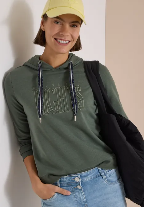 Sweatshirt mit Kapuze und Wording dried olive green Eine lächelnde Frau trägt eine grüne Kapuzenjacke, gelbe Mütze und Jeans, sie trägt eine schwarze Tasche.