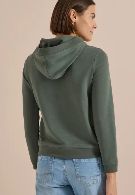Sweatshirt mit Kapuze und Wording dried olive green Frau trägt einen dunkelgrünen Kapuzenpullover, Rückenansicht, kombiniert mit blauen Jeans.