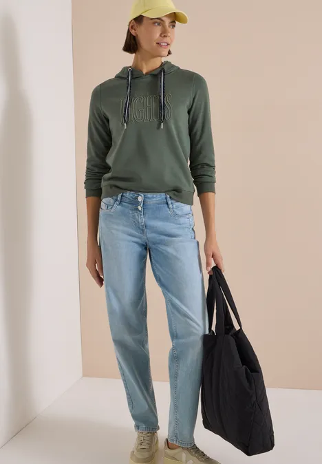 Sweatshirt mit Kapuze und Wording dried olive green Eine Frau trägt grüne Kapuzenjacke, Jeans, beige Cap und hält eine große schwarze Tasche.