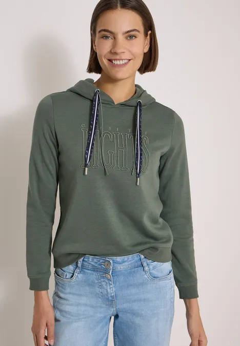 Sweatshirt mit Kapuze und Wording dried olive green Junge Frau in olivgrünem Hoodie mit Kordelzug und hellen Jeans, lächelt vor neutralem Hintergrund.