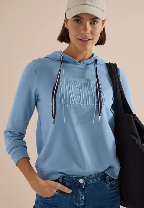 Sweatshirt mit Kapuze und Wording easy blue Frau in hellblauem Kapuzenpullover, Jeans, beiger Cap und schwarzer Tasche vor hellem Hintergrund.