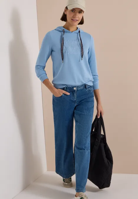 Sweatshirt mit Kapuze und Wording easy blue Frau in hellblauem Hoodie, weitem Jeans, weißer Cap und Tasche, stehend vor beigem Hintergrund