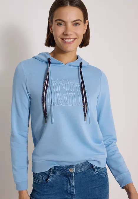 Sweatshirt mit Kapuze und Wording easy blue Frau trägt hellblauen Kapuzenpullover und Jeans, lächelt vor neutralem Hintergrund.