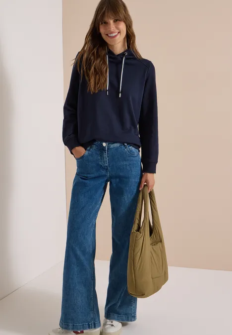 Frau trägt dunkelblauen Hoodie, weite Jeans und hält großen olivgrünen Shopper.