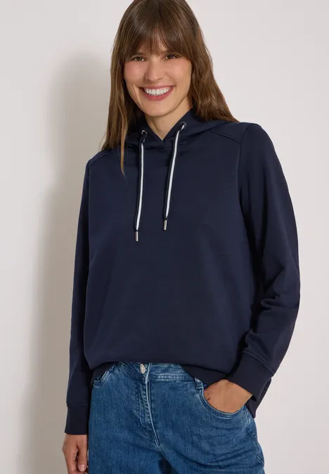 Eine Frau mit langen braunen Haaren trägt lässigen dunkelblauen Kapuzenpullover und Jeans, vor hellem Hintergrund.