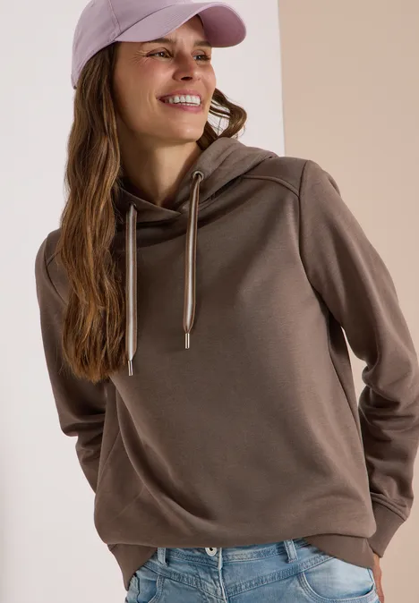 Junge Frau in Hoodie, Jeans und Baseballcap, lächelnd und seitlich blickend.