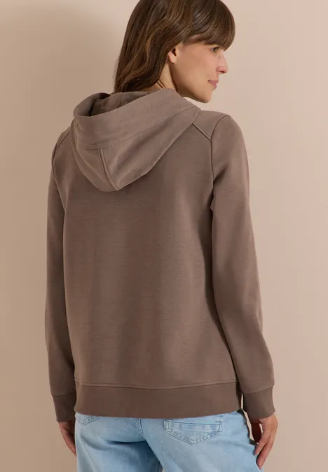 Frau von hinten in braunem Kapuzenpullover und blauen Jeans, leicht seitlich gedreht.