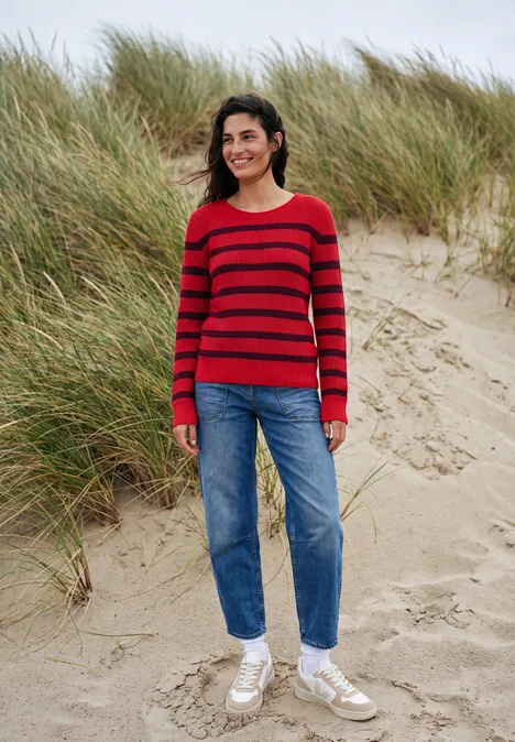 Frau mit rotem gestreiften Pullover, Jeans und weißen Sneakern steht lächelnd auf einer Sanddüne mit Gras.