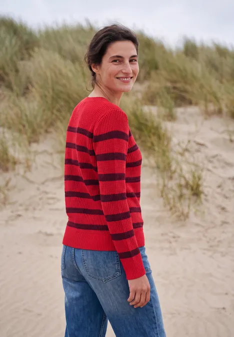 Eine lächelnde Frau in einem rot-schwarz gestreiften Pullover steht am Strand vor Dünen.