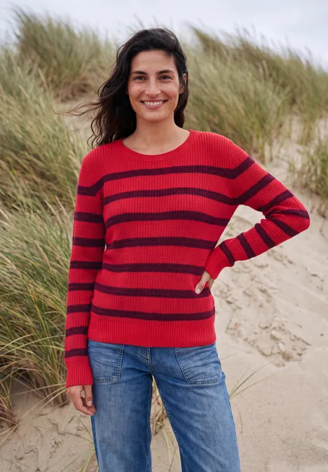 Eine lächelnde Frau in rot gestreiftem Pullover und Jeans steht vor Dünen und Strandhafer.