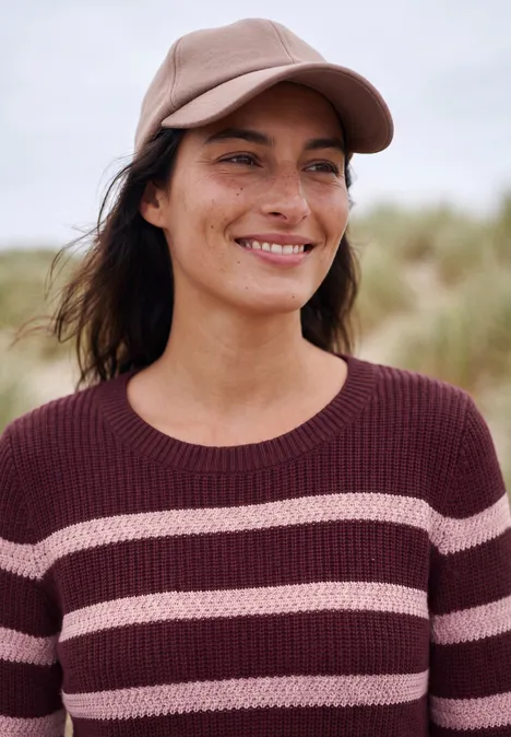 Lächelnde Frau mit Basecap und gestreiftem Pullover steht draußen vor verschwommenem Hintergrund.