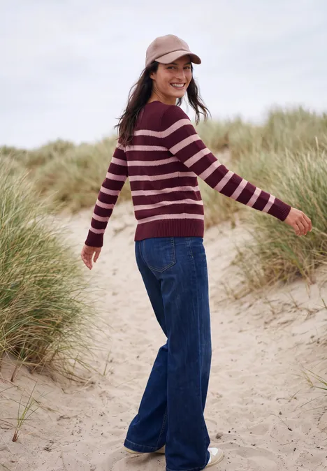 Frau in gestreiftem Pullover und Jeans spaziert lächelnd durch Dünen.