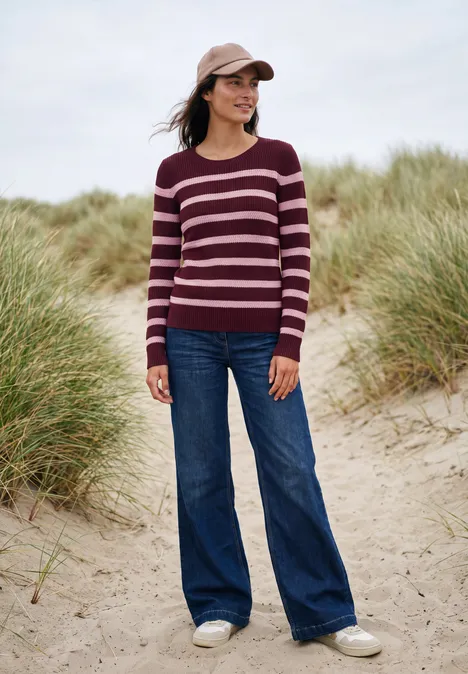 Frau mit gestreiftem Pullover, Jeans und Cap steht lächelnd in Dünenlandschaft am Strand.