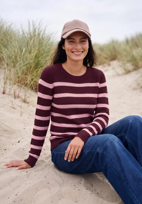 Eine lächelnde Frau sitzt am Strand in Jeans, gestreiftem Pullover und Cap, Dünen im Hintergrund.