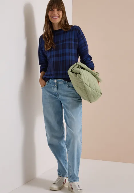 Sweter z rękawem typu dolman w kratkę urban dark blue Uśmiechnięta kobieta w niebieskim kraciastym swetrze, jasnych dżinsach i trampkach, trzyma zieloną pikowaną kurtkę.