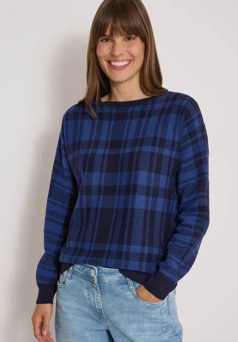 Sweter z rękawem typu dolman w kratkę urban dark blue Kobieta uśmiecha się, ma na sobie niebieski kraciasty sweter i dżinsy na neutralnym tle.