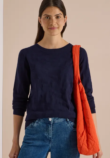 Trui met boothals en structuur urban dark blue Een vrouw met donkerblauw shirt en spijkerbroek draagt een oranje schoudertas.