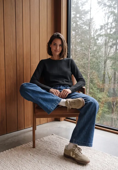 Femme assise détendue sur chaise en bois devant une grande fenêtre, en jean et pull noir, vue sur la nature.