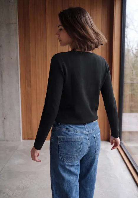 Femme de dos devant une fenêtre, portant un pull noir et un jean bleu.