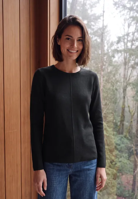 Femme souriante en pull noir et jean, debout près d'une fenêtre avec une forêt en arrière-plan.