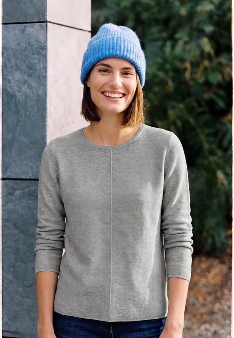 Eine junge Frau in grauem Pullover und blauer Mütze lächelt im Freien vor einer Steinwand und Grünpflanzen.